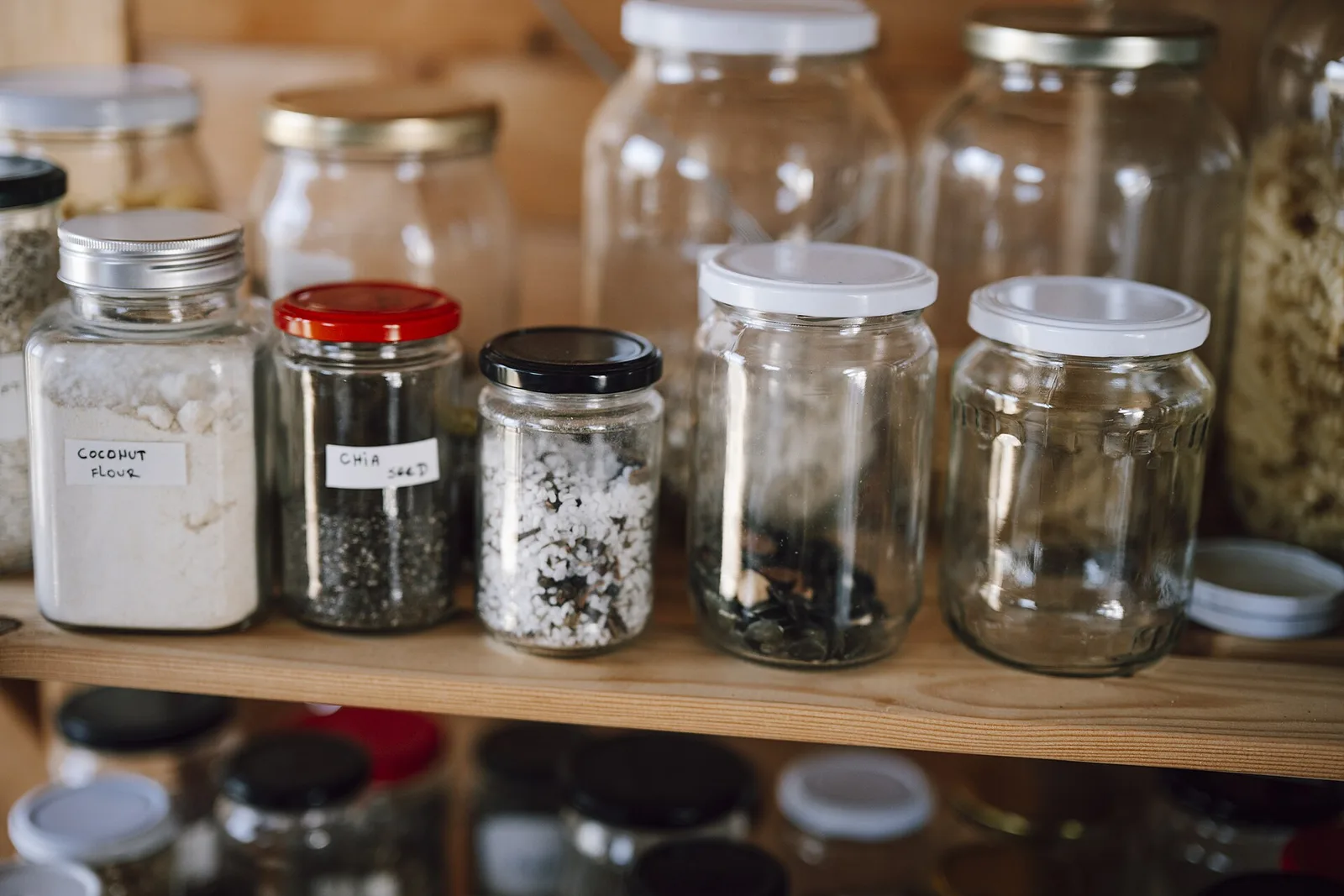 Shelving in a kitchen pantry shows various jars and bottles storing dry food items. The organized setup includes grains, seeds, and snacks. Labels help identify each container's contents.